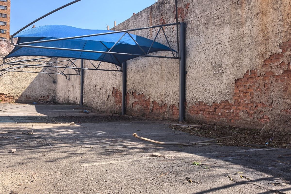Vacant commercial land featuring an asphalt parking area with a blue shade cloth carport structure and a perimeter brick wall requiring maintenance.