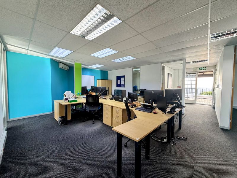 A functional open-plan office space featuring suspended ceilings, recessed lighting, air conditioning, and carpeted flooring with access to an outdoor balcony area.