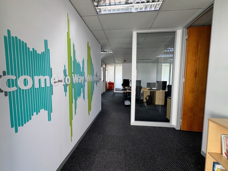 A professional office corridor featuring a glass-partitioned workspace, suspended ceiling with recessed lighting, and patterned carpet flooring.