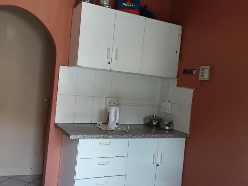 A compact kitchen area featuring white built-in cabinetry, a tiled splashback, and a stone-look laminate countertop, accessed through a classic arched doorway.