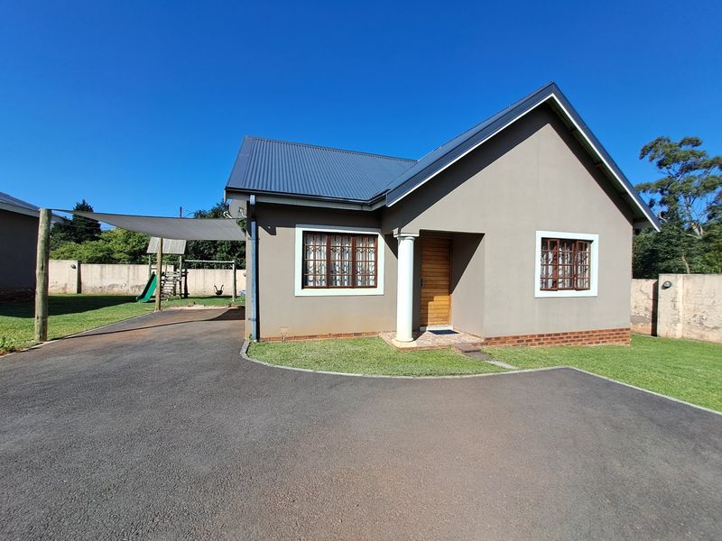 A well-maintained single-story house featuring a metal roof, asphalt driveway, and a shaded outdoor area with a neat front lawn.