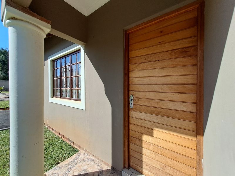 A covered entrance area featuring a solid wooden front door, a structural column, and tiled flooring overlooking a small garden space.