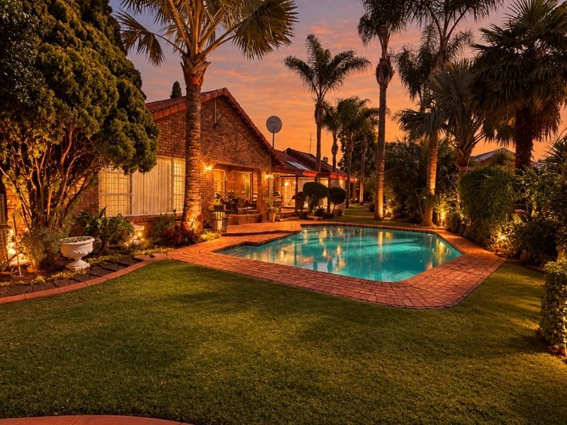 A beautifully landscaped backyard featuring a swimming pool, brick paved surround, and mature palm trees, illuminated by ambient evening lighting.