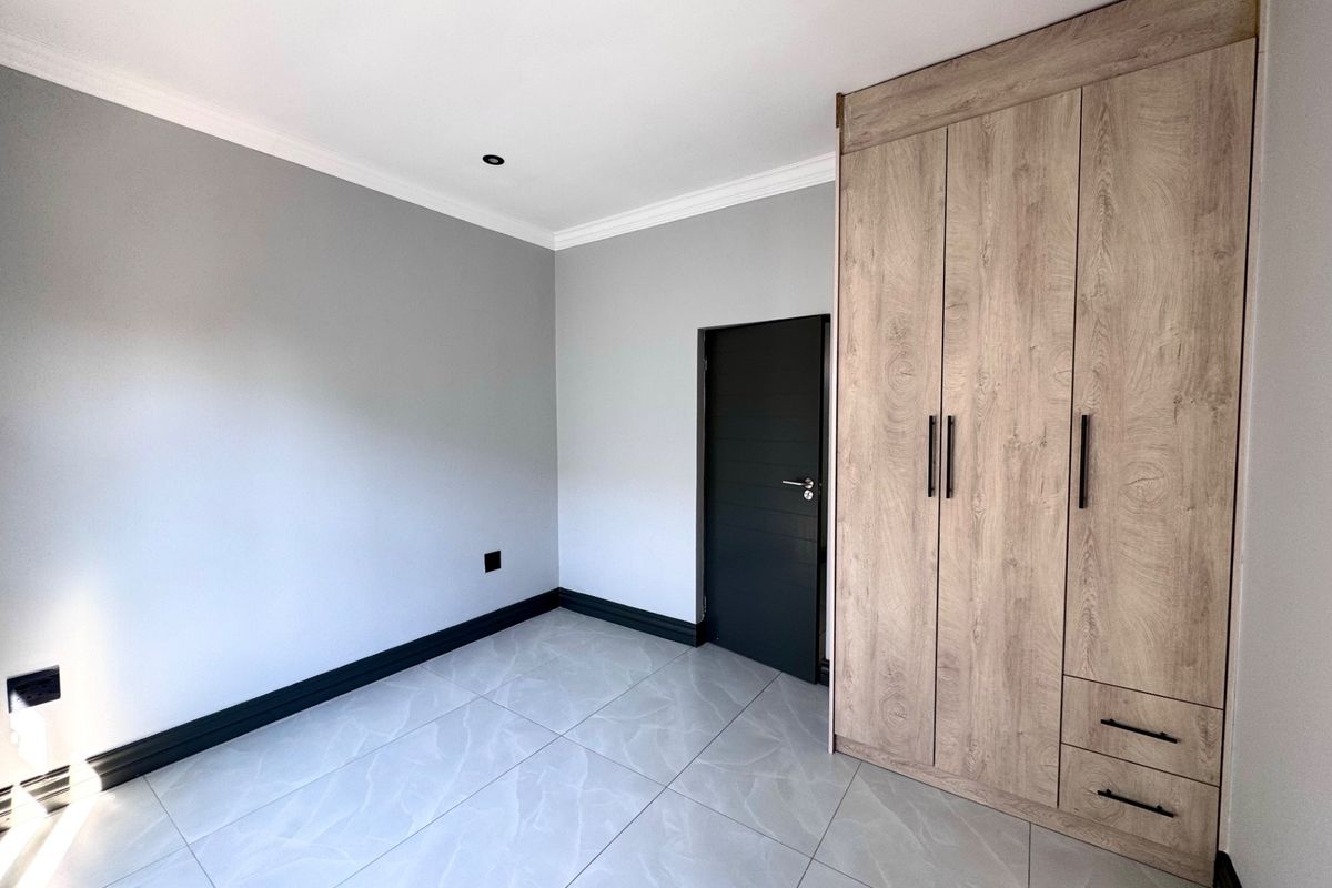 A bright, modern bedroom featuring neutral grey walls, large format floor tiles, and contemporary wood-finish built-in cupboards.