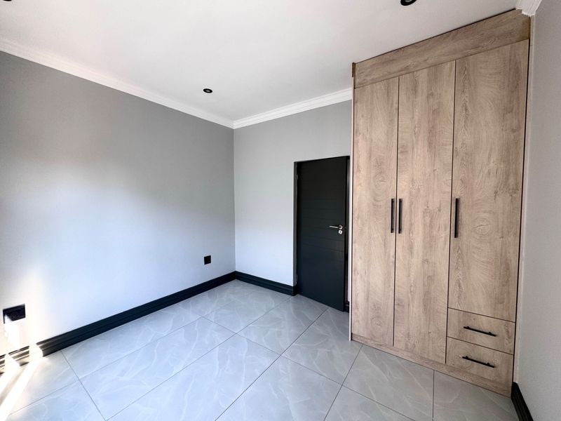 A bright, modern bedroom featuring neutral grey walls, large format floor tiles, and contemporary wood-finish built-in cupboards.