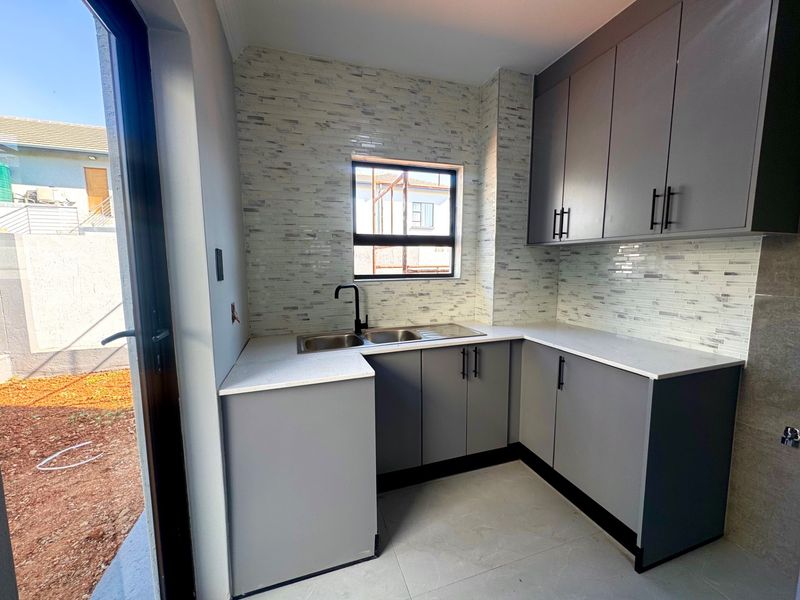 A modern, compact kitchen featuring grey cabinetry, stone countertops, a double bowl sink, and a stylish tiled backsplash with direct access to the exterior.