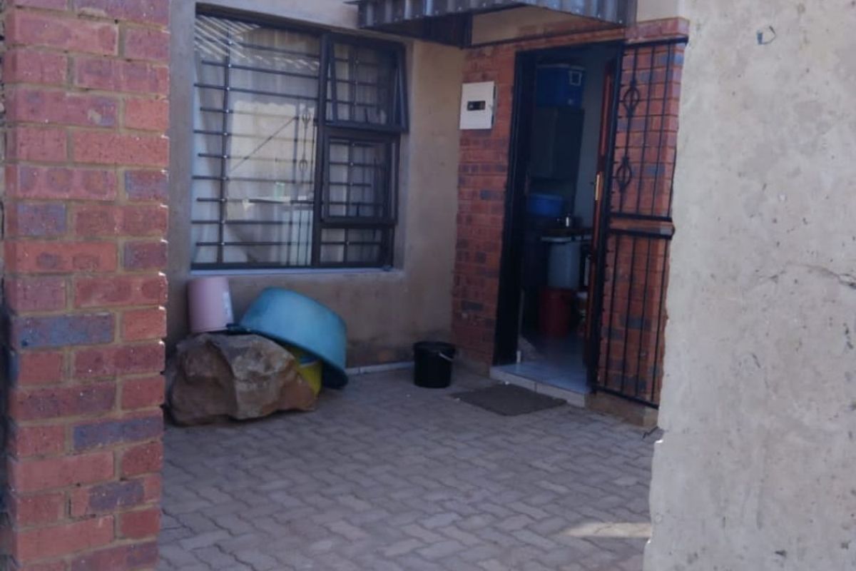 A rear exterior view showing a paved courtyard area with security bars on the window and door, and an overhead metal awning.