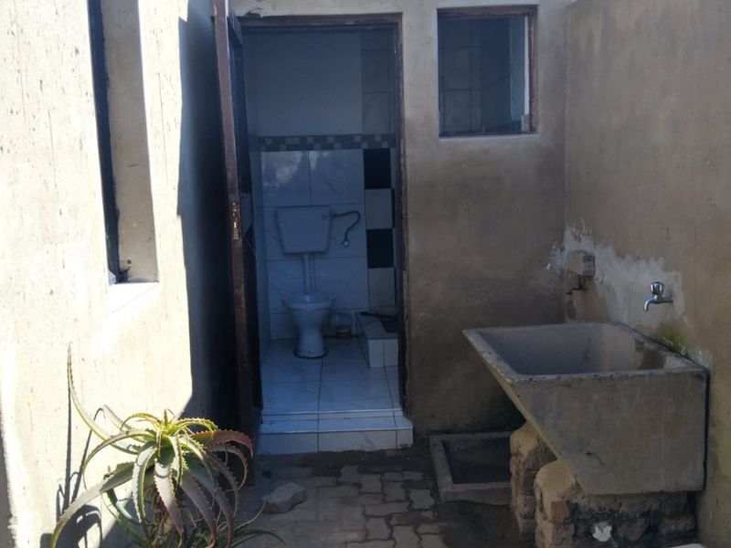 Exterior rear area featuring a concrete utility sink, brick paving, and access to an external toilet facility with weathered wall finishes.