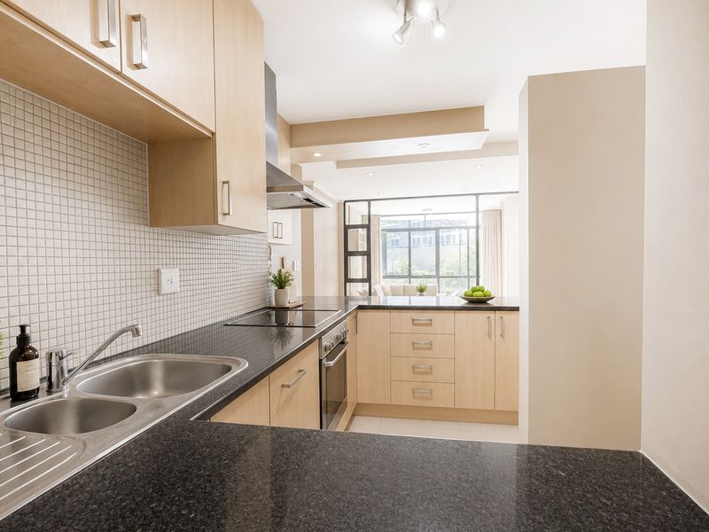 Modern kitchen featuring light wood cabinetry, dark granite countertops, mosaic tile backsplash, and integrated stainless steel appliances, flowing into an open-plan living area.