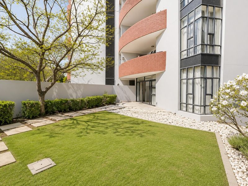 A well-maintained private garden area featuring a manicured lawn, stone pathways, and pebble landscaping, adjacent to the apartment building's rear facade and balconies.