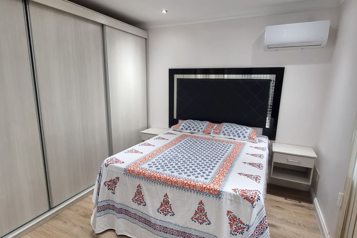 A well-maintained bedroom featuring light wood-tone laminate flooring, a large built-in wardrobe with sliding doors, and a wall-mounted air conditioning unit.
