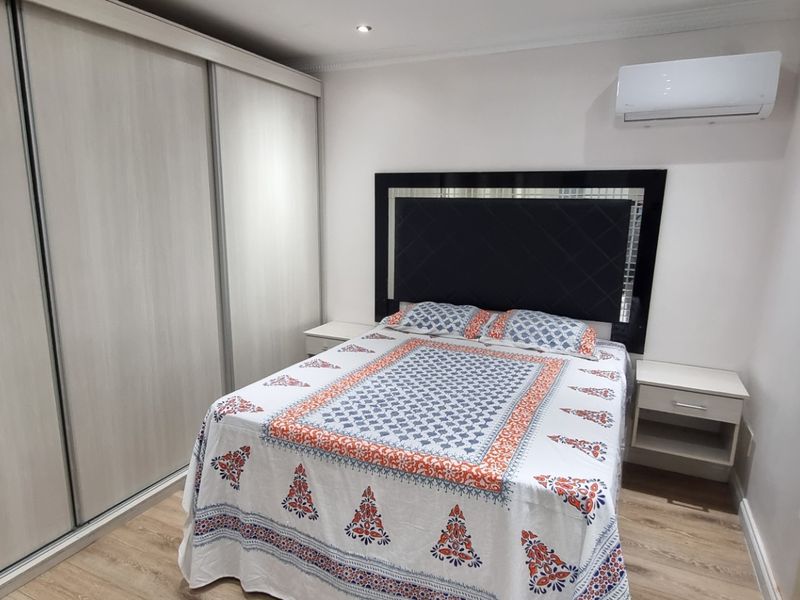 A well-maintained bedroom featuring light wood-tone laminate flooring, a large built-in wardrobe with sliding doors, and a wall-mounted air conditioning unit.