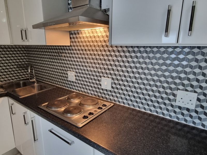 A modern, compact kitchen featuring white cabinetry, dark laminate countertops, a stainless steel hob with extractor fan, and a geometric patterned tiled backsplash.