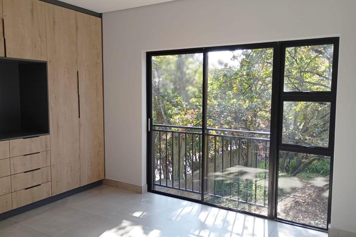 A bright, modern bedroom featuring built-in wooden cupboards, large sliding glass doors, and neutral tiled flooring with a view of the surrounding greenery.