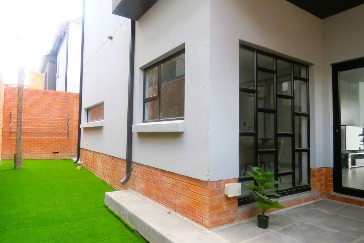 A modern rear exterior view featuring a combination of brick and plastered walls, aluminium window frames, a paved patio area, and artificial lawn.
