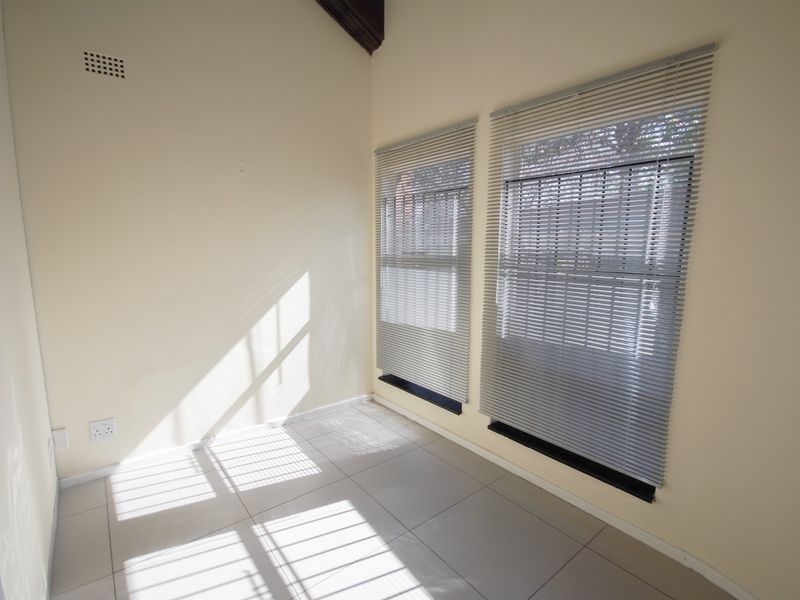 A bright, empty office space featuring tiled flooring, a vaulted ceiling with an exposed timber beam, and large windows fitted with venetian blinds.
