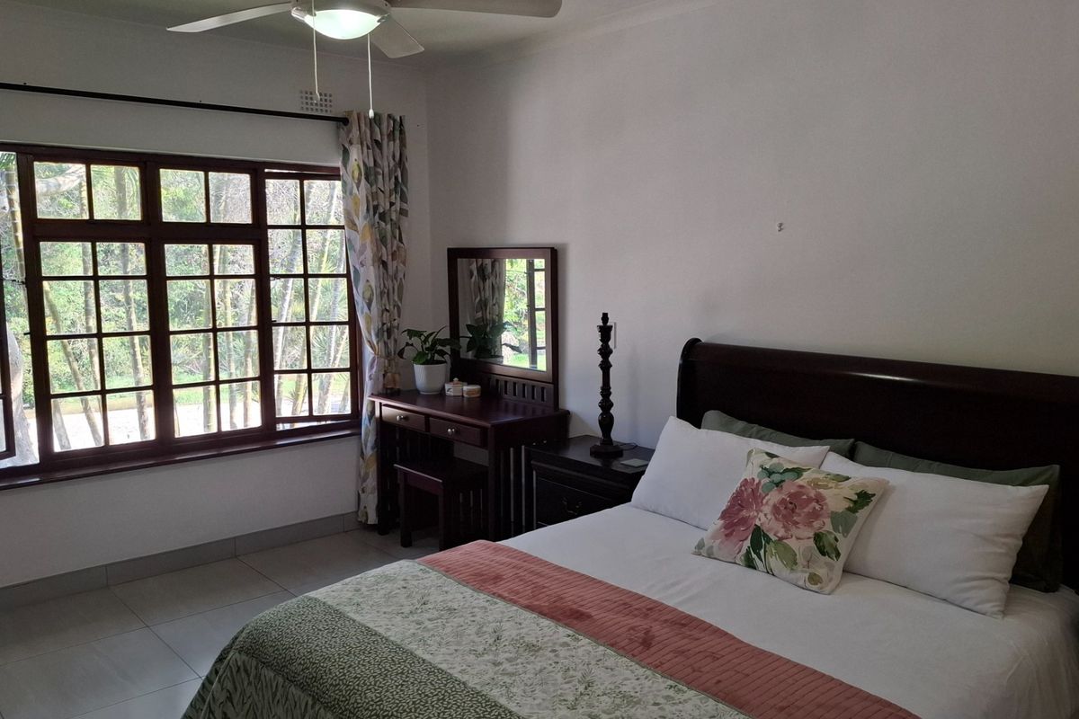 A well-lit bedroom featuring tiled flooring, a ceiling fan, and large wooden-framed windows with security bars overlooking the garden.