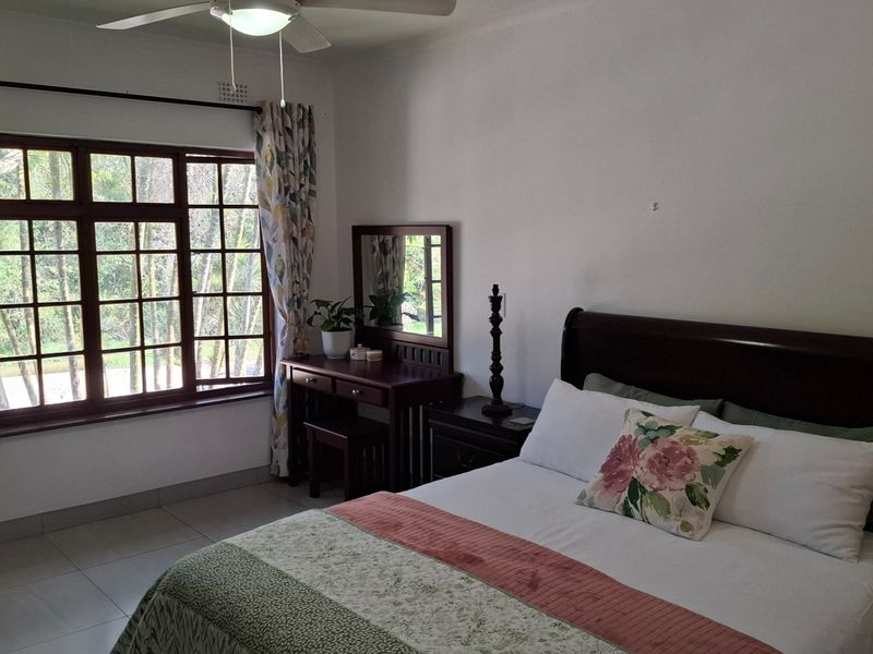 A well-lit bedroom featuring tiled flooring, a ceiling fan, and large wooden-framed windows with security bars overlooking the garden.