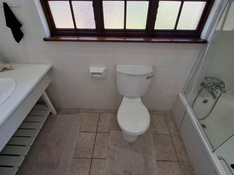 A functional bathroom featuring a built-in vanity, toilet, and a shower-over-bath combination with tiled finishes and a timber-framed window.
