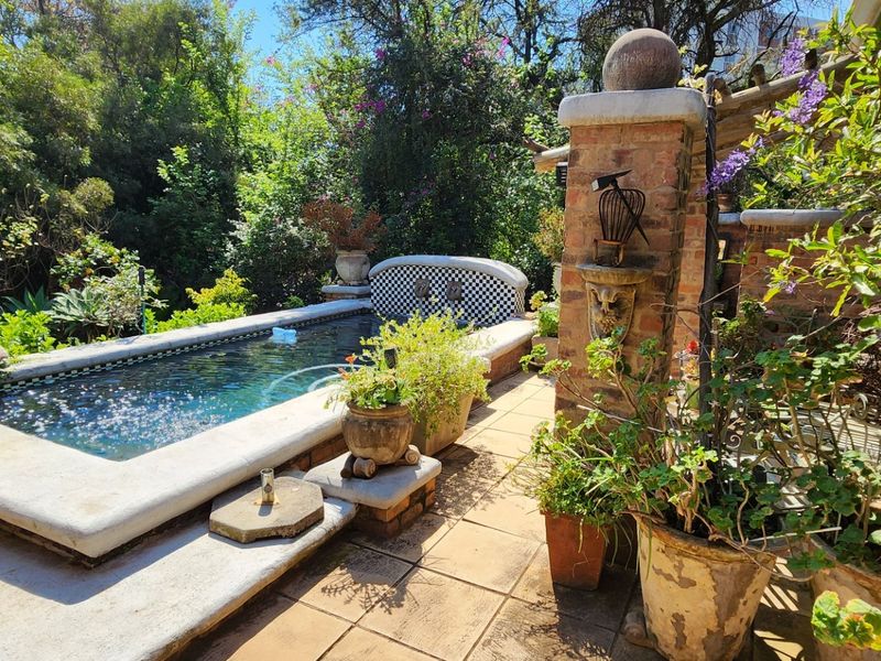 A private outdoor pool area featuring a tiled patio, brick pillar water feature, and lush surrounding garden landscaping.