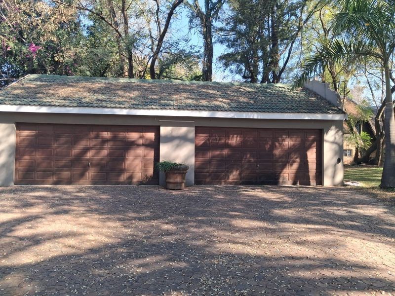 A detached double garage featuring brown sectional doors and a tiled roof, situated on a paved driveway surrounded by mature trees.