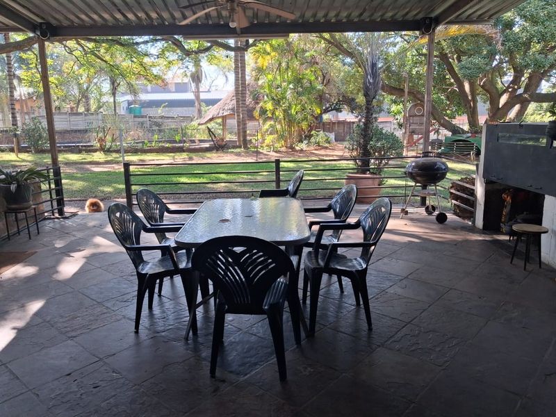 A spacious covered patio featuring tiled flooring, a built-in braai, and a ceiling fan, overlooking a large garden area.