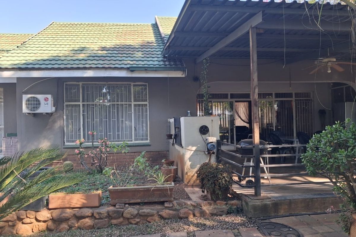 Rear view of the property featuring a covered patio, built-in braai, and established garden area with stone retaining wall.
