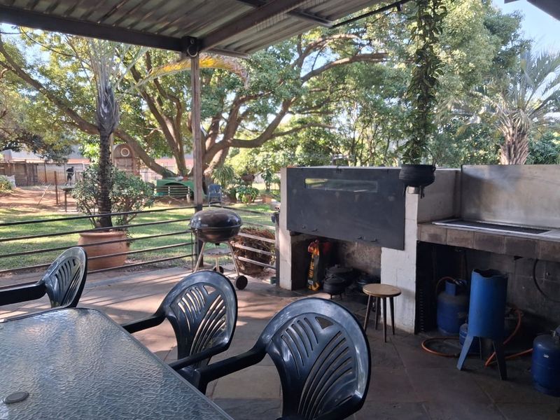 A covered outdoor patio area featuring a built-in braai and tiled flooring, overlooking a spacious garden with mature trees.