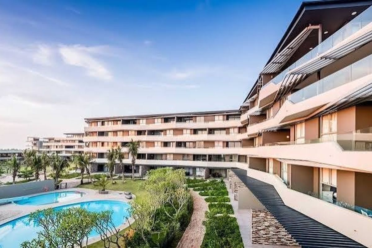 A wide-angle view of a modern apartment complex featuring tiered balconies, glass railings, landscaped gardens, and a communal swimming pool area.