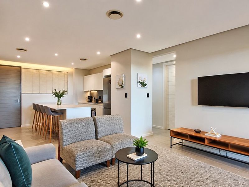 Modern open-plan living area featuring a sleek kitchen with island, tiled floors, recessed lighting, and a wall-mounted television in a well-maintained apartment setting.