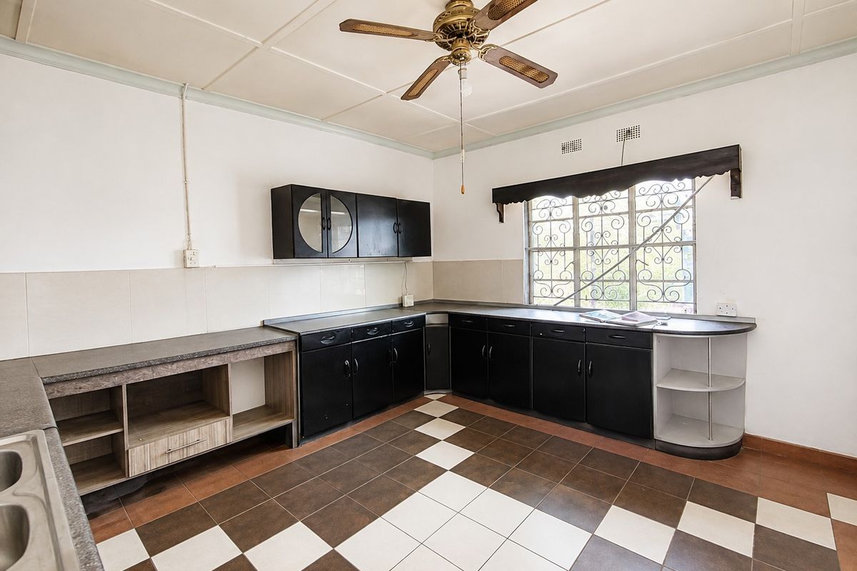 A spacious, empty kitchen featuring dark built-in cabinetry, laminate countertops, a ceiling fan, and a checkerboard tiled floor with security bars on the window.