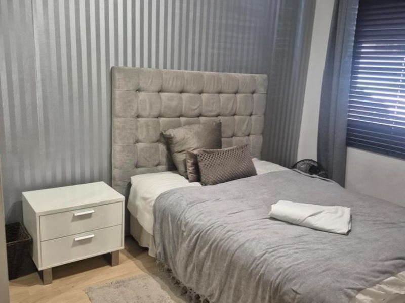 A bedroom featuring laminate flooring, a feature striped wallpaper wall, and window blinds.