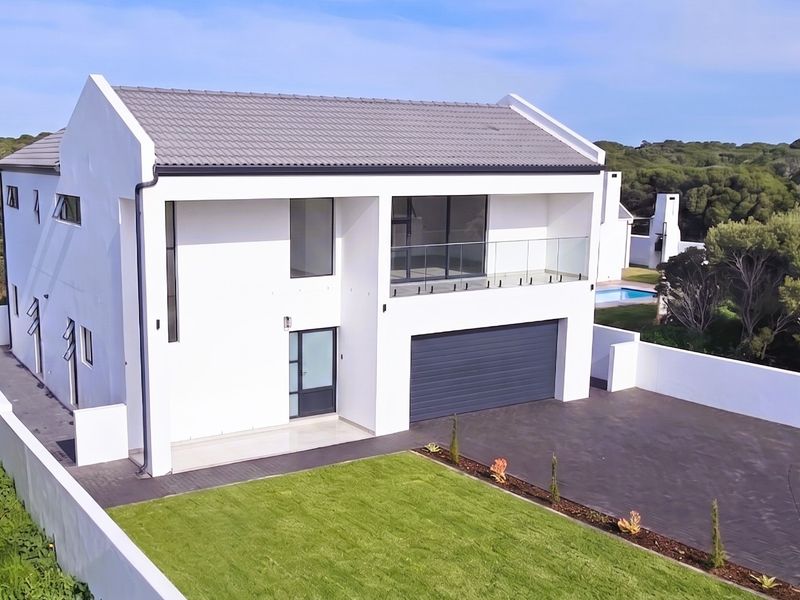 Modern double-story residence featuring a clean white facade, tiled roof, glass balcony, paved driveway, and an integrated garage, set within a landscaped property.