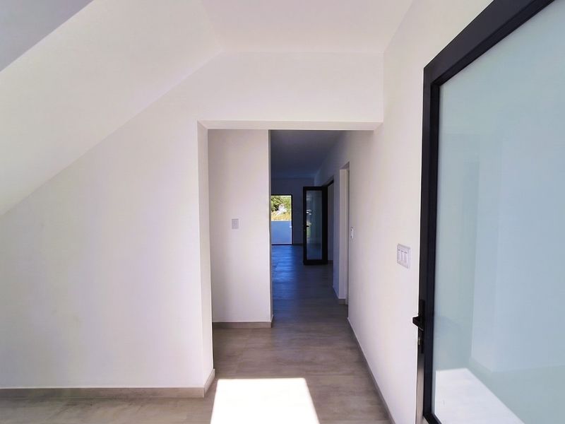 A bright, modern hallway featuring neutral tiled flooring and clean white walls, leading through to an open-plan living area with glass door access.