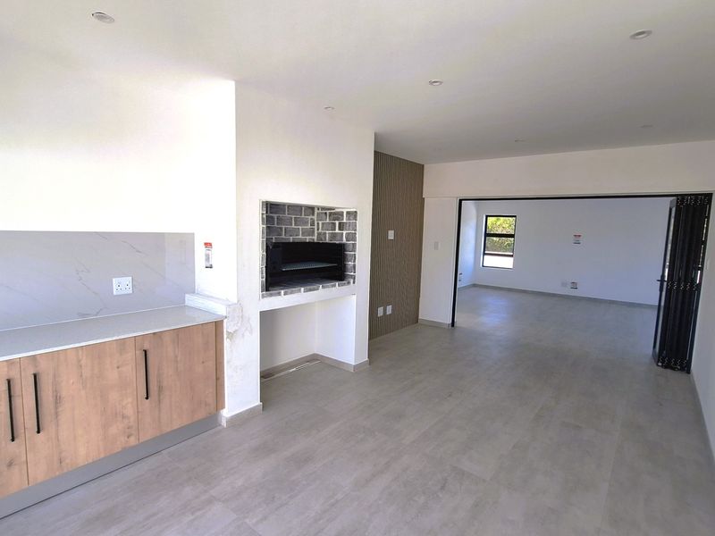 Modern open-plan living area featuring a built-in braai, sleek kitchen cabinetry with stone countertops, and neutral flooring, leading to an adjacent room via stacking doors.
