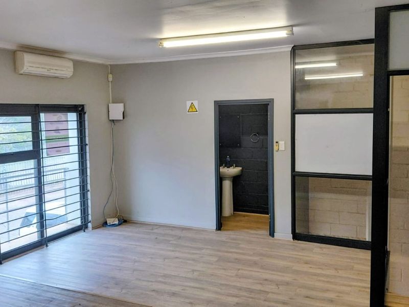 A clean, open-plan office space featuring laminate flooring, a glass partition, security bars on the window, and an integrated air conditioning unit.