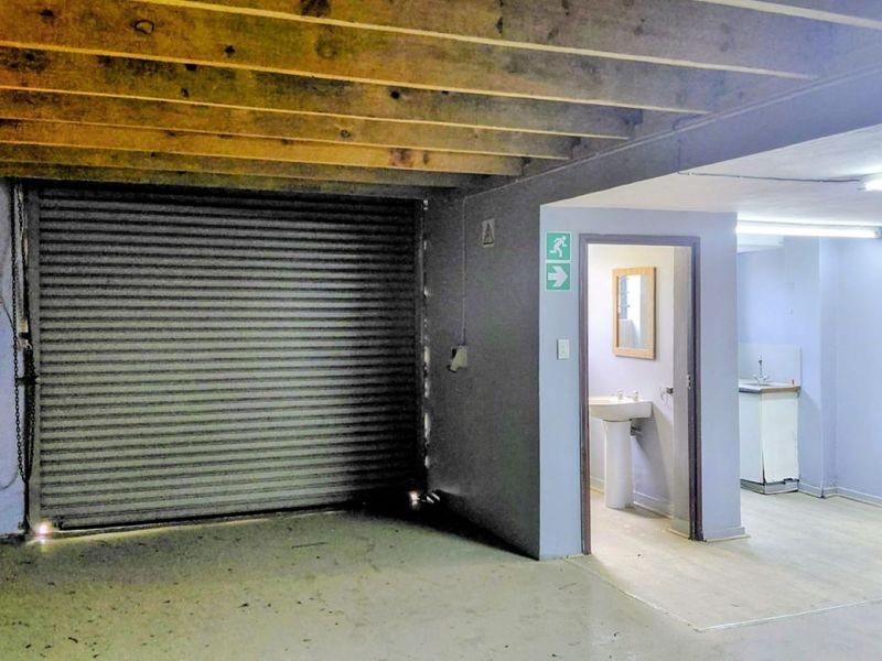 Industrial garage space featuring a roller shutter door, exposed timber ceiling joists, and an adjoining washroom area with a pedestal basin.
