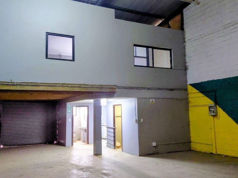 Industrial warehouse space featuring a roller shutter door, mezzanine level, and integrated utility rooms with exposed structural elements and painted block walls.