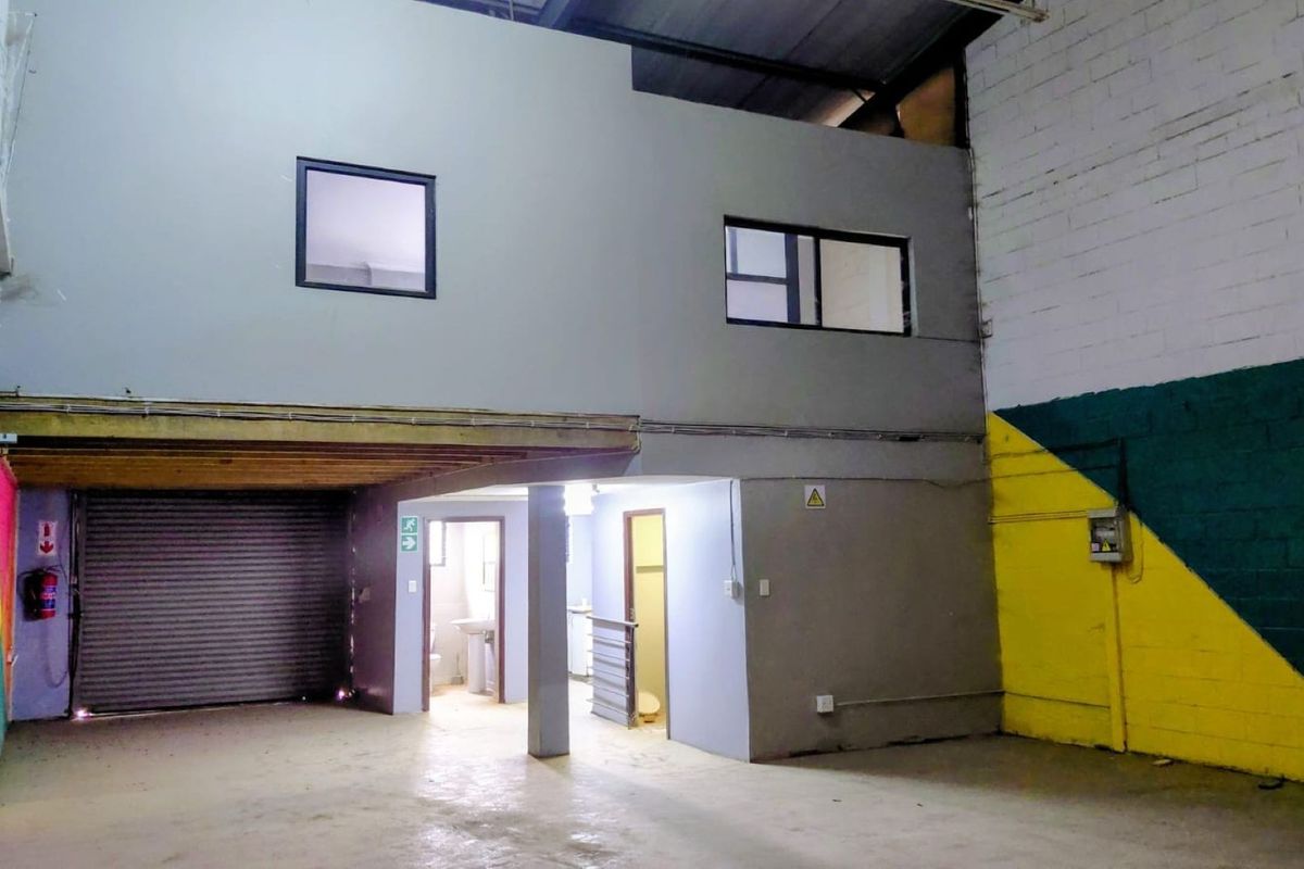 Industrial warehouse space featuring a roller shutter door, mezzanine level, and integrated utility rooms with exposed structural elements and painted block walls.