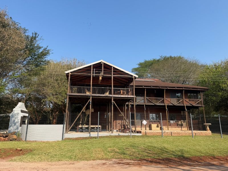 A two-story timber-clad house featuring a large raised deck, balcony, and an outdoor brick braai area set within a fenced garden.