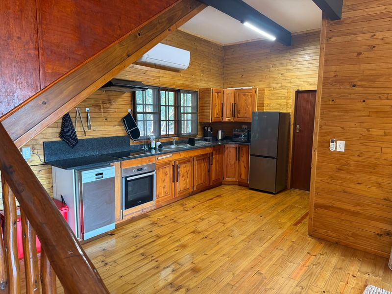 A rustic kitchen featuring timber wall cladding, wooden cabinetry, granite countertops, and integrated appliances, situated beneath an exposed timber staircase.