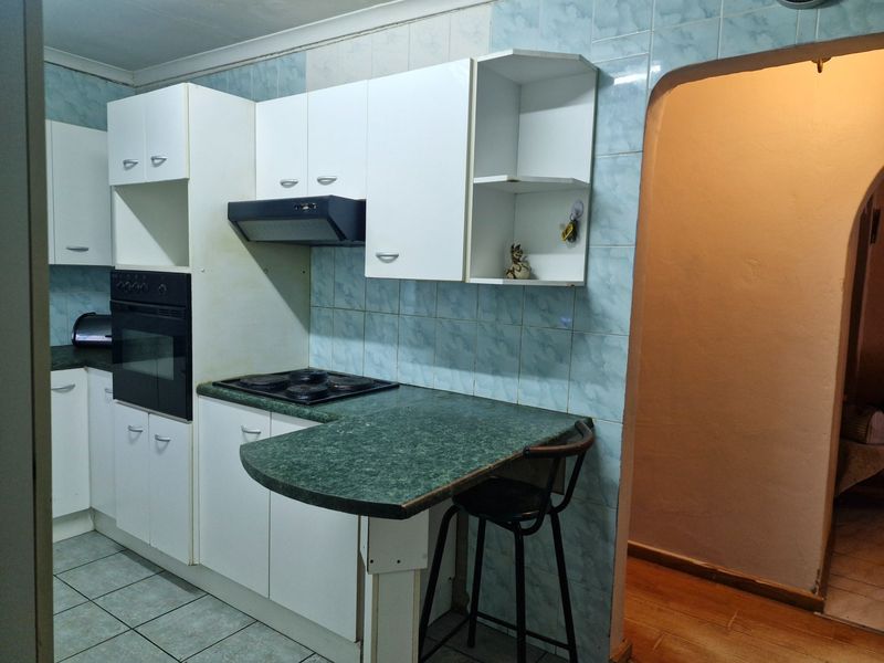 A compact kitchen featuring white built-in cabinetry, a green laminate countertop with breakfast bar, built-in oven, electric hob, and blue tiled backsplash.
