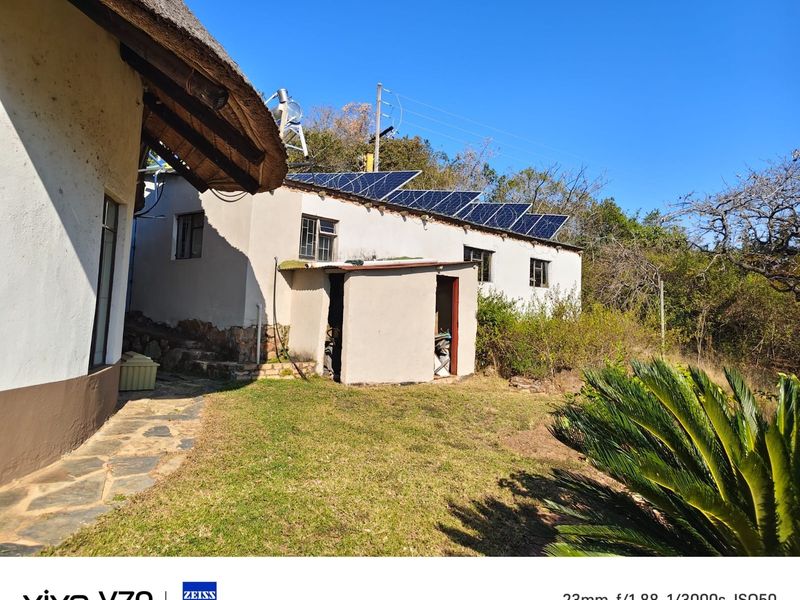 Rear view of the farm property featuring a traditional thatch roof, solar panel installation, and a stone paved walkway leading to a utility shed.