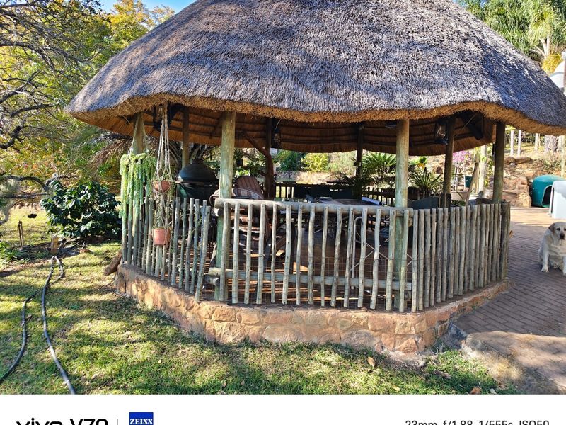 A rustic outdoor thatched lapa area featuring a timber pole structure, stone base, and paved flooring, ideal for garden entertainment.