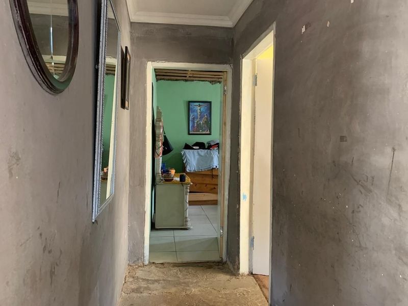 A narrow hallway featuring unfinished plaster walls and exposed flooring, leading into an adjacent bedroom space.