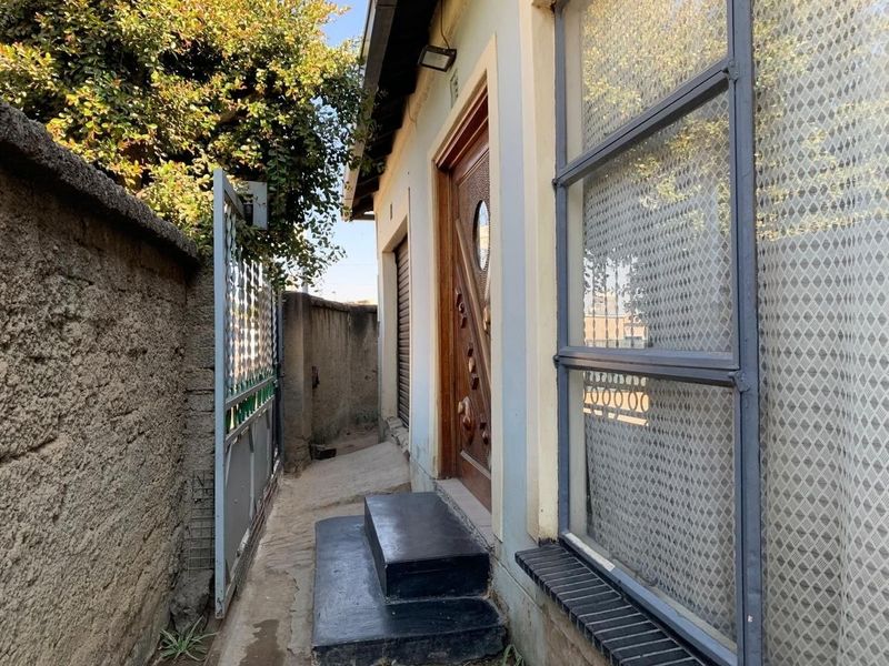 Side entrance area featuring a solid wood door, steel-framed windows, and concrete steps, enclosed by a perimeter wall and security gate.