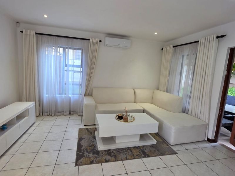 A bright and well-maintained living room featuring neutral tiled flooring, recessed downlighting, and a wall-mounted air conditioning unit.