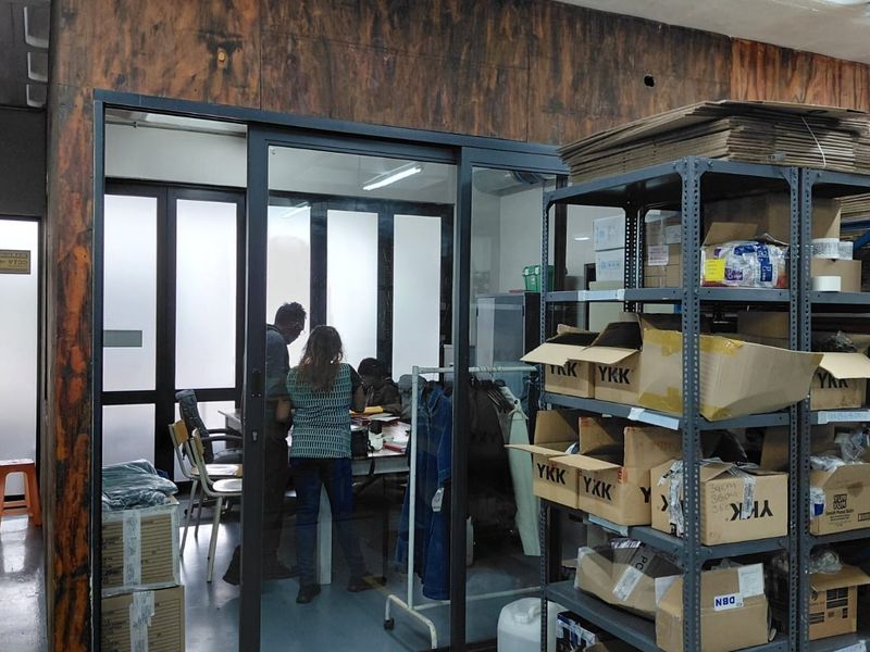 Industrial office space featuring a glass-partitioned private office area, exposed ceiling ducting, and heavy-duty metal shelving units for storage.