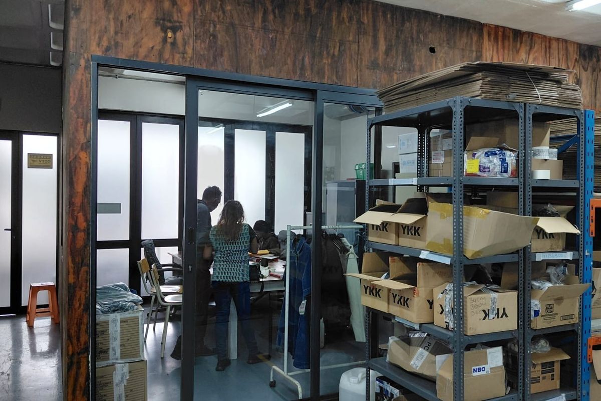 Industrial office space featuring a glass-partitioned private office area, exposed ceiling ducting, and heavy-duty metal shelving units for storage.