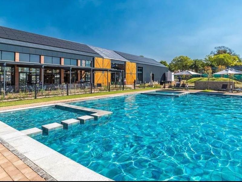 A well-maintained outdoor swimming pool area featuring modern paving, metal fencing, and a large clubhouse building with solar panels in the background.